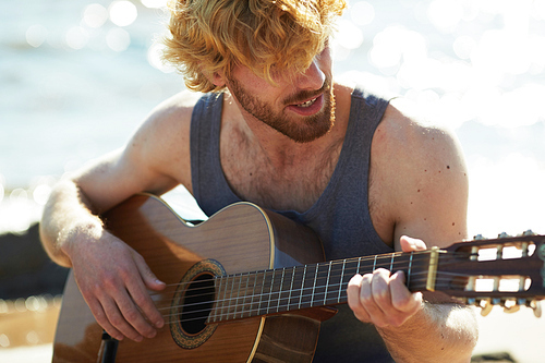 Happy guy in grey vest playing guitar by seaside on sunny summer day