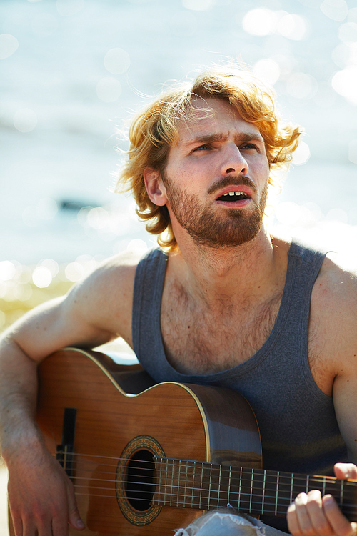 Young man with wavy hair singing while playing his guitar on the beach during vacation