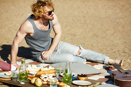 Young man in sunglasses, vest and jeans sitting on sand by served place with snack