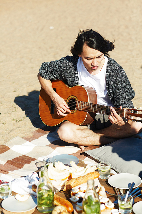 Young man playing guitar while sitting on sand with snack near by