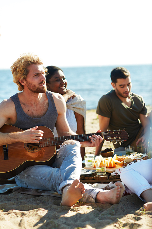 Guy with guitar and his friends sitting on sand, relaxing and having snack on summer day