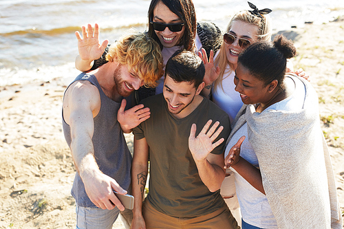 Cheerful friends waving hands while recording video or making selfie in smartphone while enjoying summer day on the beach