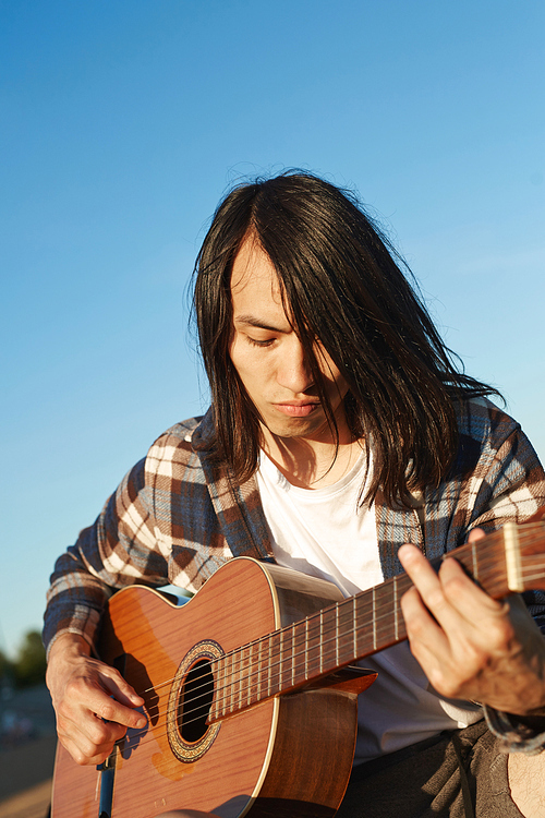 Young man with dark long hair playing guitar against blue sky on summer day