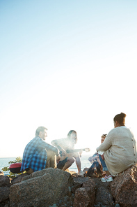 Young people sitting on huge stones around campfire on summer evening