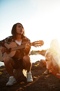 Guy with guitar sitting on stone and singing for his friends on summer evening by campfire