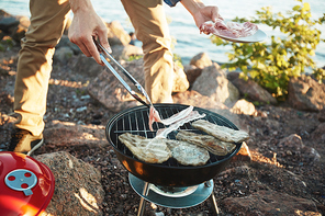 Young man frying bacon and meat on grill during picnic by seaside