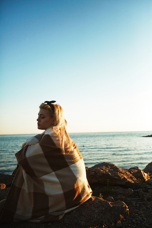 Young woman wrapped in woolen plaid sitting on stone by seaside at sunset