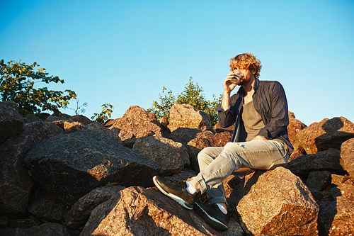 Young adventurer sitting on stones and having refreshing drink on summer evening after hot day