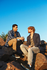 Two friendly guys with drinks sitting on huge stones and having chat on hot summer day