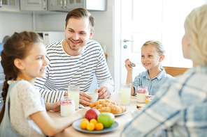 Young family of four sitting by served table at breakfast and having talk in the kitchen