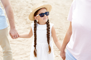 Portrait of cute little girl holding hands with mom and dad while enjoying walk along beach in Summer