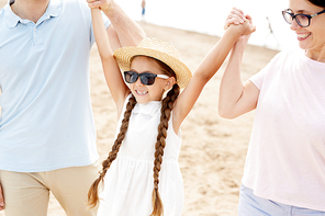 Portrait of happy little girl holding hands with mom and dad while enjoying walk along beach during Summer vacation