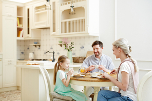 Happy excited young family in casual clothing having breakfast together: they laughing and talking while eating pancakes at one table in domestic kitchen