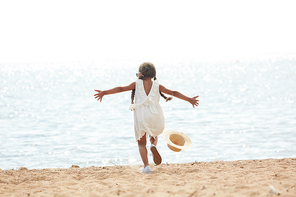 Little girl in white dress running after her blown hat towards water while trying to catch it