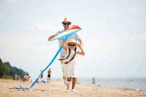 Adorable girl in white dress, hat and sunglasses and her father running down sandy beach while playing with kite