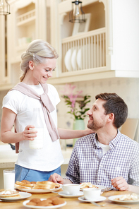 Happy loving attractive young wife with wrapped sweater around neck taking care of husband and holding bottle of milk while talking to beloved man in domestic kitchen