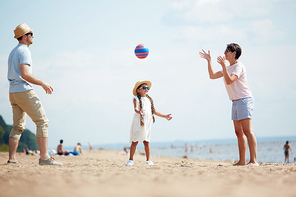 Young family of three standing on sand in circle and throwing ball to each other while playing by seaside