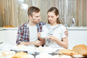 Young man embracing his wife with cup of tea during talk by dinner table in the kitchen