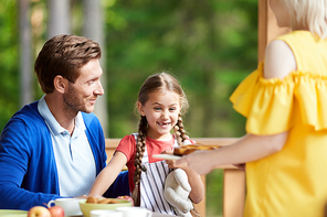 Cheerful girl looking at homemade apple pie held by her mom while sitting by table with father