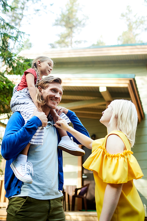 Happy young man with little daughter on his shoulders standing in front of wife while enjoying weekend in the country