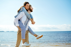 Laughing young woman embracing man while sitting on his back and enjoying summer day by seaside