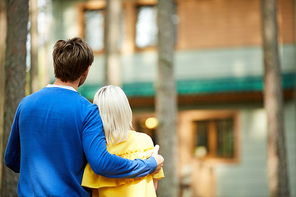 Back view of young affectionate embracing couple standing in front of their country house