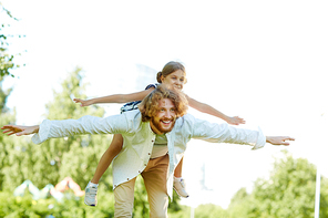 Cute little girl on her father back and the man stretching arms while having fun on summer weekend in park