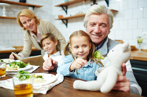 Happy mature man holding teddybear while his granddaughter giving food to her toy by dinner