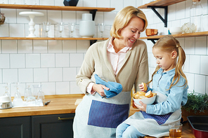 Happy mature female and her little granddaughter with bowl of wheat bread sitting in the kitchen