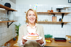 Smiling woman in birthday cap holding self-made cake for her guests