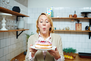 Mid-aged woman blowing burning candles on birthday cake in the kitchen