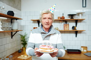 Mature grey-haired man in birthday cap holding cake with candles during home celebration