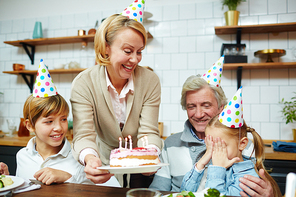 Little girl covering eyes by hands while her grandmother with birthday cake looking at her by festive dinner