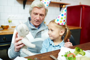 Granddaughter in birthday cap looking at teddybear held by her grandfather while playing by table