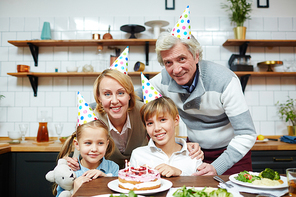 Adorable kids and their grandparents in birthday caps sitting by festive table