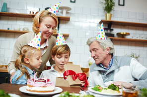 Little boy in birthday cap looking at his present in giftbox with his sister and grandparents near by