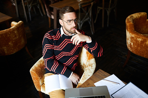 Pensive man with open notebook sitting in beige velvet armchair by table and thinking of organization ideas