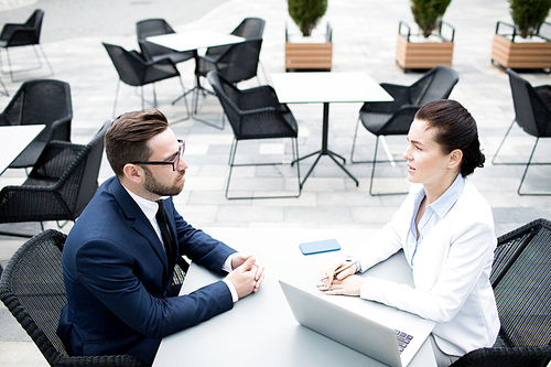 From above side view of coworking elegant man in black and woman in white sitting with laptop at table and talking