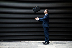 Side view of young successful male in business dress, eyeglasses throwing briefcase near black profiled sheeting