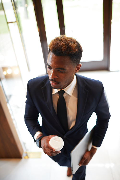 Young African businessman in suit holding coffee and laptop walking into an office building