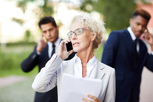 Serious confident mature lady lawyer with curly hair wearing eyeglasses calling client on mobile phone while viewing papers, businessmen talking by phones in background