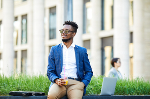 Happy healthy African-American businessman in smart casual outfit drinking juice from disposable mug and sitting on concrete slab while looking around