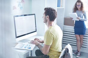 Young designer working in front of computer monitor by desk with his colleague reading papers on background