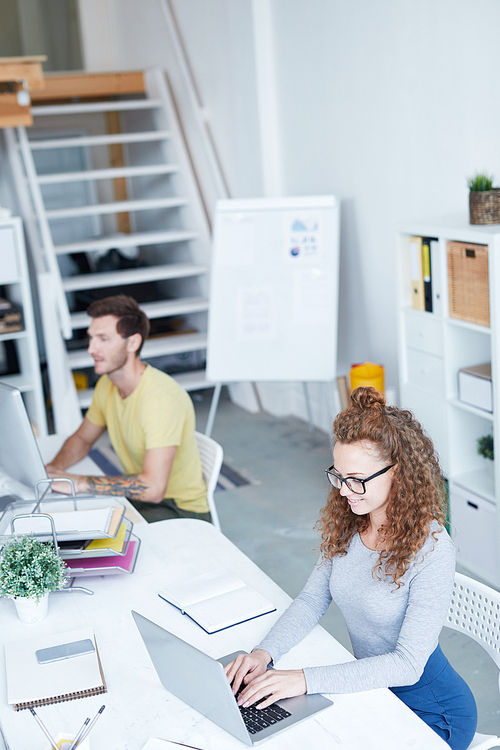 Young woman sitting by desk in front of laptop and typing with her colleague networking near by