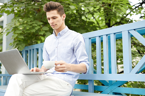 Young man with cup of coffee sitting on bench in summer cafe with laptop in front and searching for online ideas