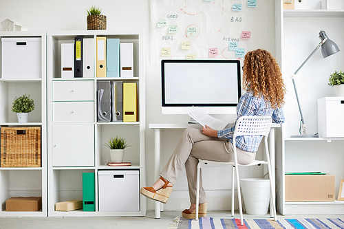 Young designer with red curly hair sitting on chair by desk in front of computer monitor during work