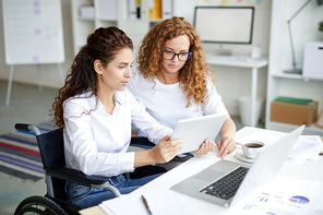 Young woman in wheelchair and her colleague discussing online data by desk in office