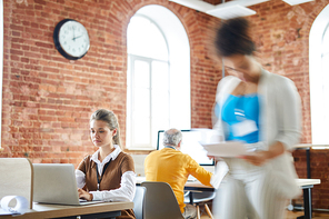 Young serious office worker browsing in the net while sitting in front of laptop with colleague networking behind and blurry woman walking near by