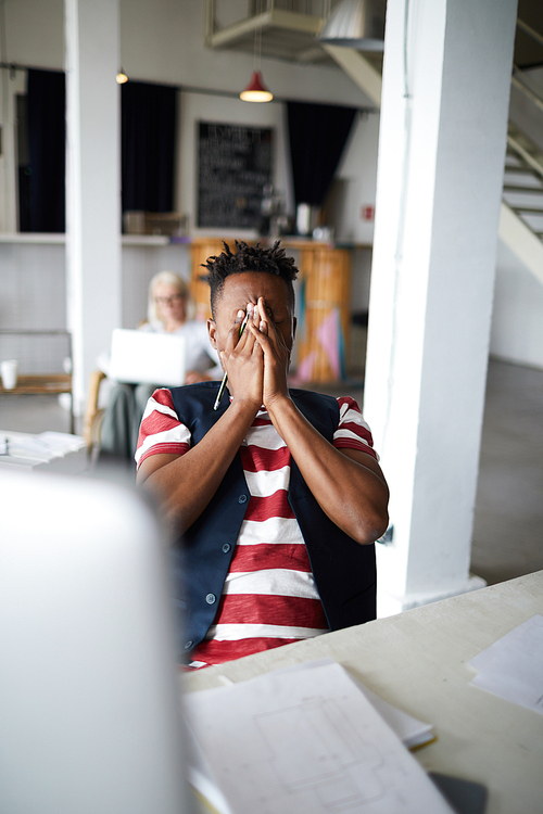 Tired or frustrated businessman hiding his face in hands while sitting by workplace in office