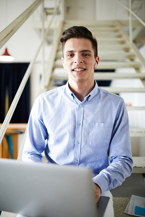 Young confident man in light blue shirt sitting on staircase in office, networking and looking at you